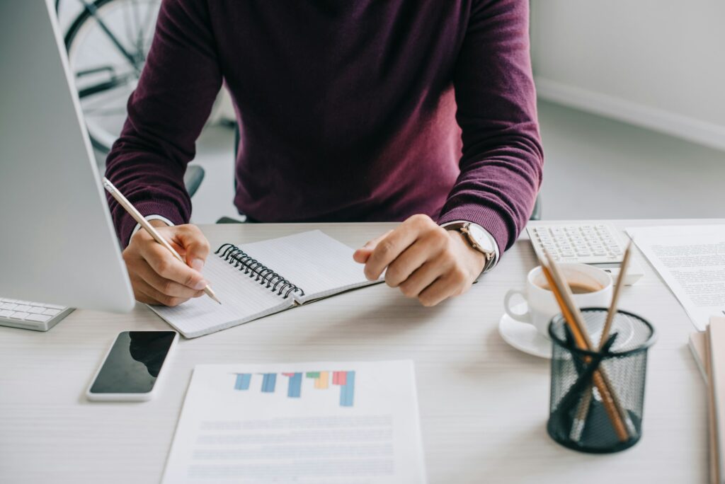 cropped image of designer in burgundy sweater writing something to notebook in office